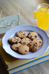 Plate of chocolate chip cookies, stack of books, reading glasses, orange soda, mobile phone and pen on the table. Hygge at home. Pastel colors, selective focus.