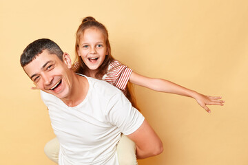 father and daughter wearing casual t-shirts standing isolated over beige background man carrying child on his back small kid sitting piggyback on her daddy.