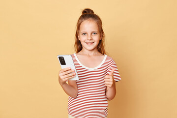 Cute optimistic smiling little brown haired girl wearing striped t-shirt standing isolated over beige background holding smartphone browsing internet showing thumb up.