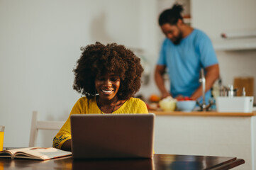 A happy african american female entrepreneur is working from home on a laptop while her husband is cooking.