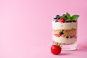 Trifle dessert in a glass with berries, mint, whipped cream and biscuit on a pink background....