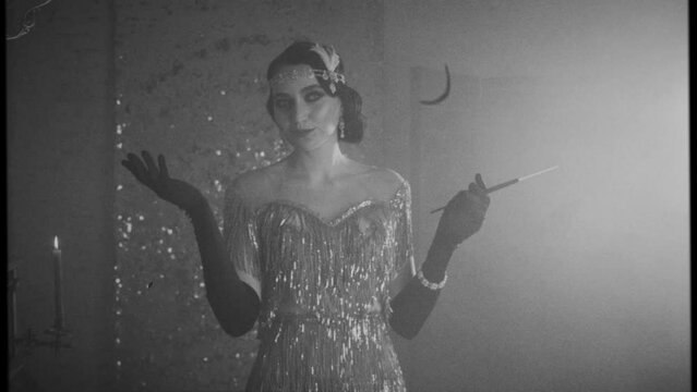 Antique stylistics black white film added noise. Mysterious smile charming young beautiful retro woman 20s. Headband hairstyle finger wave. Long gloves mouthpiece cigarette. Sparks smoke. Silver dress
