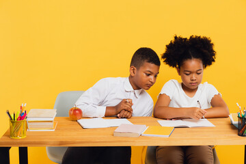 Happy african american schoolgirl and schoolboy sitting together at desk and studying on yellow background. Back to school concept