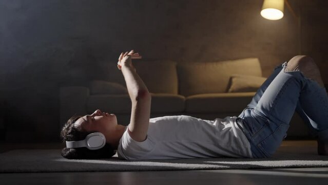 Teenager boy listening relaxing music on headphones lying on the floor at night. Meditation and mindfulness concept.
