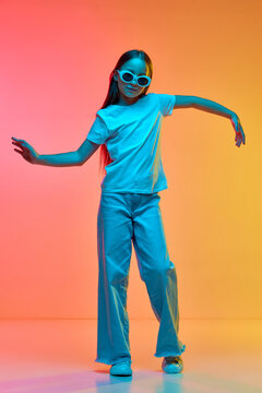 Portrait of cute school age girl, child wearing casual style clothes dancig over neon light background. Concept of childhood, music, fun