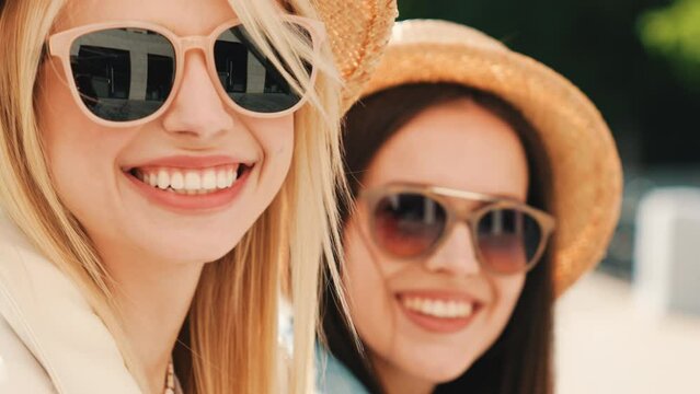 Two young beautiful smiling hipster women in trendy summer clothes. Carefree models posing on street background. Positive models having fun. Cheerful and happy, at sunny day. In hat and sunglasses