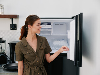 Woman smiling with teeth looking into camera in kitchen at home opened freezer empty with ice inside, home refrigerator, defrosted, view from back.