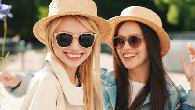 Two young beautiful smiling hipster women in trendy summer clothes. Carefree models posing on street background. Positive models having fun. Cheerful and happy, at sunny day. In hat and sunglasses