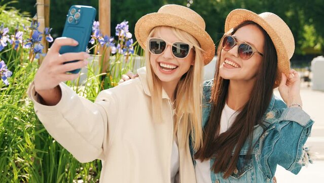 Two young beautiful smiling hipster women in trendy summer clothes. Carefree models posing on street background. Positive models having fun. Cheerful and happy. In hat and sunglasses. Taking selfie