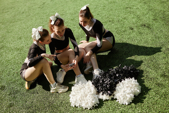 High angle view of young cheerleading girls sitting on green grass and using smartphone, they resting after training