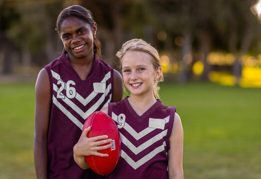 two school-aged girls in football jerseys holding aussie rules football
