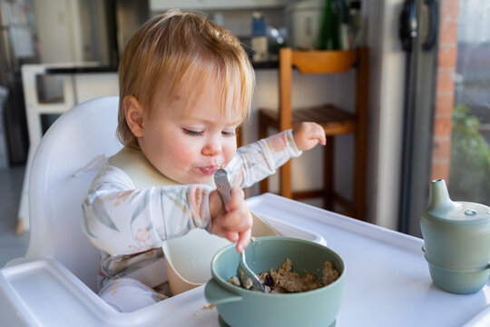 One year old baby girl using spoon to eat weetbix and sultanas for breakfast