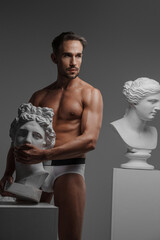 A seductive male model dressed only in underwear, posing with an ancient Greek bust sculpture pressed against his body, set against a gray background