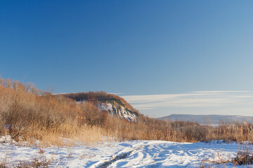 Winter landscape. Road covered with snow and a hill with tall dry grass on a sunny day.
