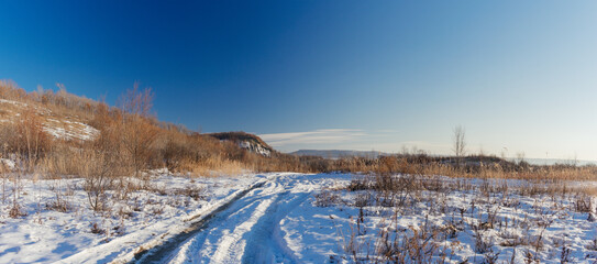 Winter landscape. Road covered with snow and a hill with tall dry grass on a sunny day.