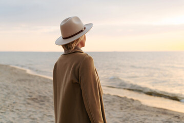 Back view of lonely pensive woman in coat and hat standing on seashore at sunset in evening. Stylish mature lady looking at sea and horizon