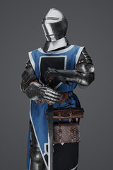 Fototapeta premium Medieval guard dressed in armor and blue surcoat holding a sword, standing in statue pose, against a gray background