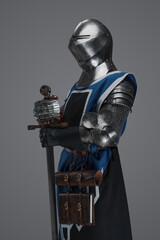 A medieval soldier garbed in blue surcoat and armor holding a sword with a still, statue-like pose,...