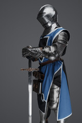 Naklejka premium A medieval soldier garbed in blue surcoat and armor holding a sword with a still, statue-like pose, against a gray background