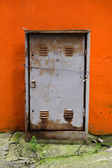 Old gray metal rusty grunge door to an electrical switchboard against a bright orange house wall, abstract background