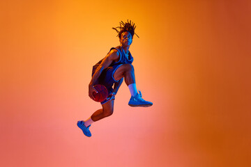 Dynamic image of athletic young man, basketball player in motion, jumping with ball against orange background in neon lights. Concept of professional sport, competition, hobby, game, competition, ad