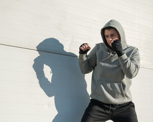 A man in a hoody trains boxing against a gray wall. 
