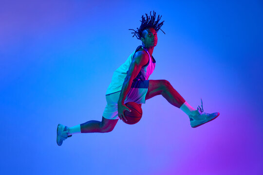Slam dunk Ambitious young man, basketball player in minions, playing basketball against gradient blue background in neon lights. Concept of professional sport, competition, hobby, competition, ad