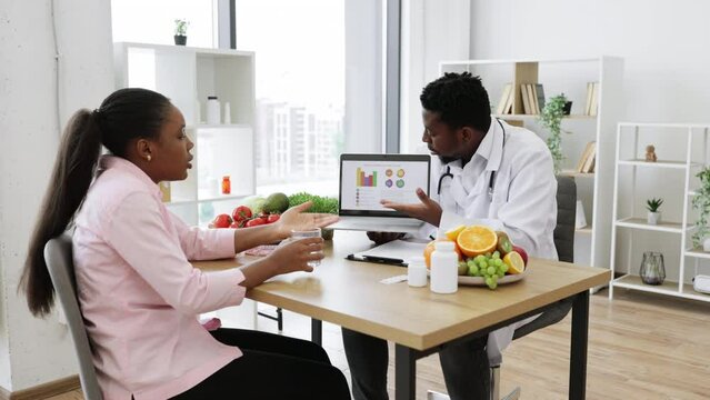 African american doctor and patient having appointment in hospital cabinet and discussing woman's treatment. Long haired lady holding glass of water while male specialist pointing at graphs on laptop.