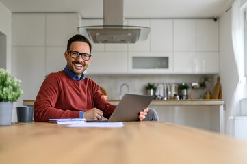 Fototapeta premium Cheerful handsome man with laptop and note pad on desk working from home office, smiling at camera