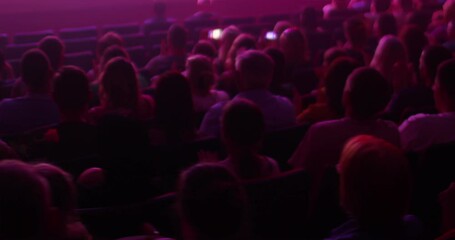 Rear view of people sitting in a full hall and cheering. Indoors. There is a dim purple light in the hall. The concept of holding events, concerts, holidays.