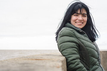 young latina woman standing leaning against a wall by the water smiling looking at the camera.