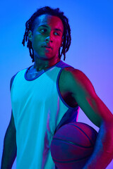 Young man with dreads, basketball player in uniforma posing with ball against gradient blue...