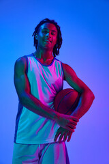 Young man with dreads, basketball player in uniforma posing with ball against gradient blue...