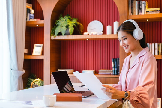 Happy Asian Woman Student Looking At Paper While Watching Webinar Or Doing Video Chat By Webcam.