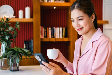 Young Asian business woman  drinking coffee using smartphone in home office. Happy smiling female professional working holding mobile phone using smartphone texting messages on cellphone.