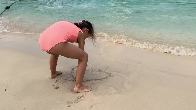 Child Playing in Sea Sand Beach, Little Girl on Tropical Exotic Sea Coastline. The girl draws a heart on a sandy beach.
