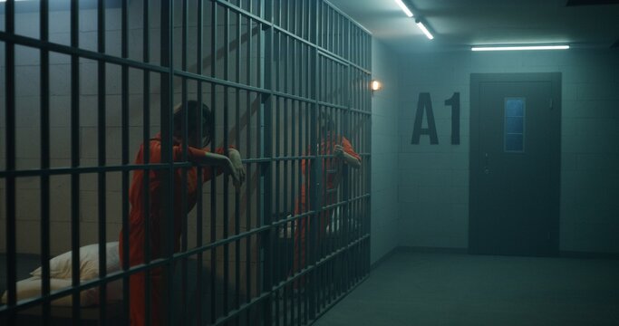 One female prisoner in orange uniform stands behind metal bars, another sits on the bed in prison cell. Women serve imprisonment terms for crimes in jail. Depressed inmates in correctional facility.