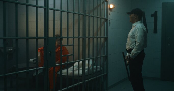 Prison officer walks the corridor, gives phone to female prisoner in orange uniform through metal bars in prison cell. Women serve imprisonment term in jail. Detention center or correctional facility.