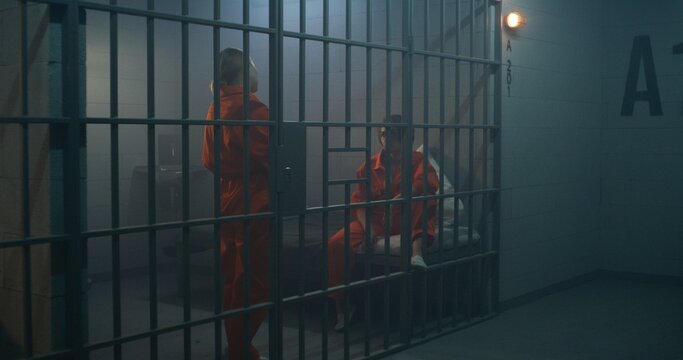 Two female prisoners in orange uniforms talk. One woman sits on the bed, another walks in prison cell. Women serve imprisonment terms for crimes in jail, detention center or correctional facility.