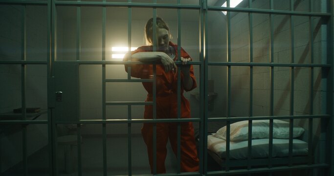 Female prisoner in orange uniform walks and stands behind metal bars, looks at barred window in jail cell. Woman serves imprisonment term for crime in prison. Depressed criminal in detention center.