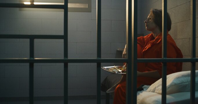 Prisoner in orange uniform gives dinner from food serving trolley to female criminal in prison cell. Woman criminal sits on the bed, serves imprisonment term for crime in jail or detention center.