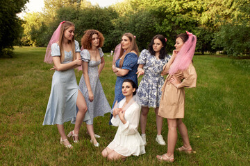 Fototapeta premium Happy charming bride in white dress with white veil sitting on the grass and bridesmaids with pink veils standing around her. Cheerful bachelorette party in a summer park.