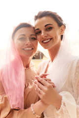 Close up portrait of happy charming bride in a white dress with a white veil and a bridesmaid in a beige dress with pink veil gently hugging and smiling. Cheerful bachelorette party in a summer park.