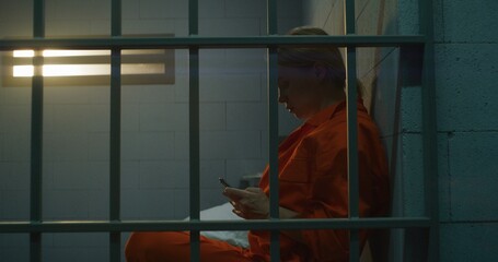 Female prisoner using smartphone in prison. Woman criminal in orange uniform makes call, sits on bed in prison cell. Serving imprisonment term in jail. Detention center or correctional facility.