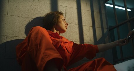 Jailer gives phone to female prisoner. Woman criminal in orange uniform makes call, sits on bed in...