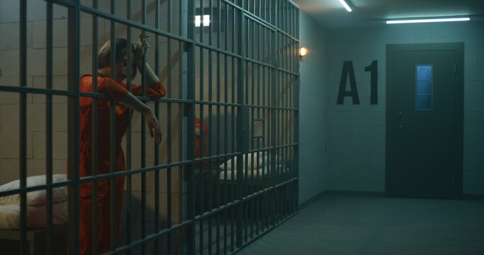 One female prisoner in orange uniform stands behind metal bars, another sits on the bed in prison cell. Women serve imprisonment terms for crimes in jail. Depressed inmates in detention center.