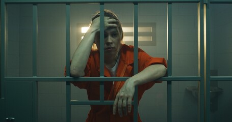 Female prisoner in orange uniform holds hands on metal bars, walks, looks at barred window in jail...