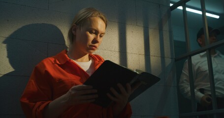 Female prisoner in orange uniform sits on bed in prison cell, reads Bible. Prison officer walks the...