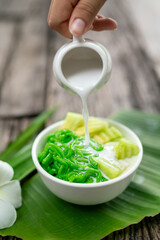 Close up of Appetizing Lod Chong being poured with fresh coconut milk on banana leaf on wooden background.