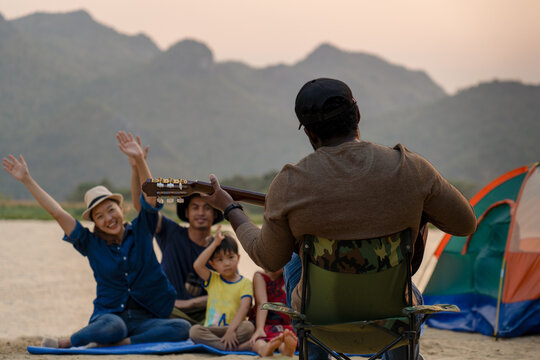 happy family with kids enjoy singing songs playing music outdoor camping area with the mountain and river beauty in nature, diverse friends travelers spend time together during holiday vacation trip